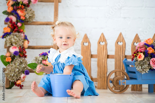Small toddler lovely girl in blue dress with a bucket and a cart of flowers in studio scenery beautiful garden. Little gardener