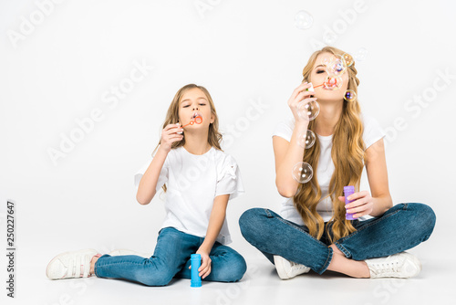 cute child with pretty mother blowing soap bubbles while sitting on floor on white background