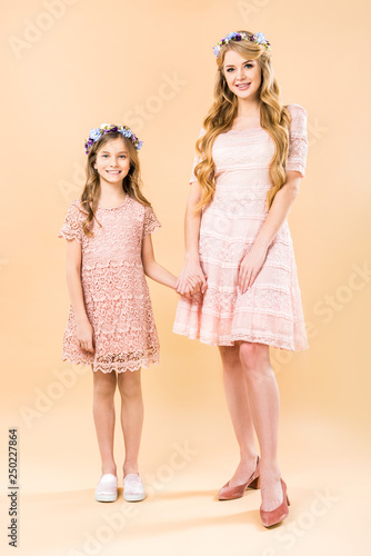 beautiful woman with adorable daughter in elegant lacy dresses and floral wreaths holding hands on yellow background photo