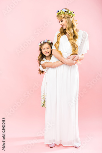 beautiful mother and adorable daughter in elegant white dresses and colorful floral wreaths on pink background