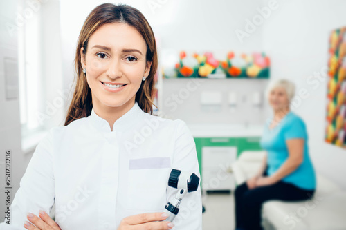 Doctor dermatologist with dermatoscope, smiling while looking at camera. Behind her sitting senior patient.