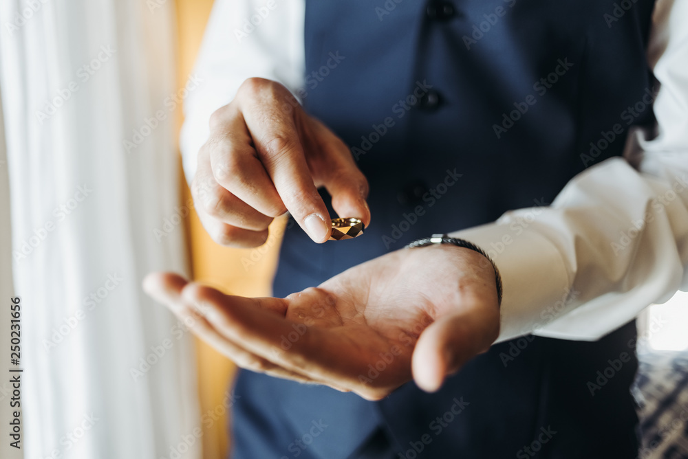 Groom holds wedding ring standing before the window in a hotel room