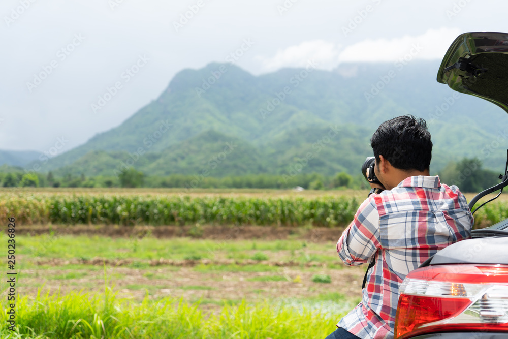 Young man sits in the car trunk take photo with dslr camera with green corn field and mountain nature background. Summer vocation with car on the road concept.