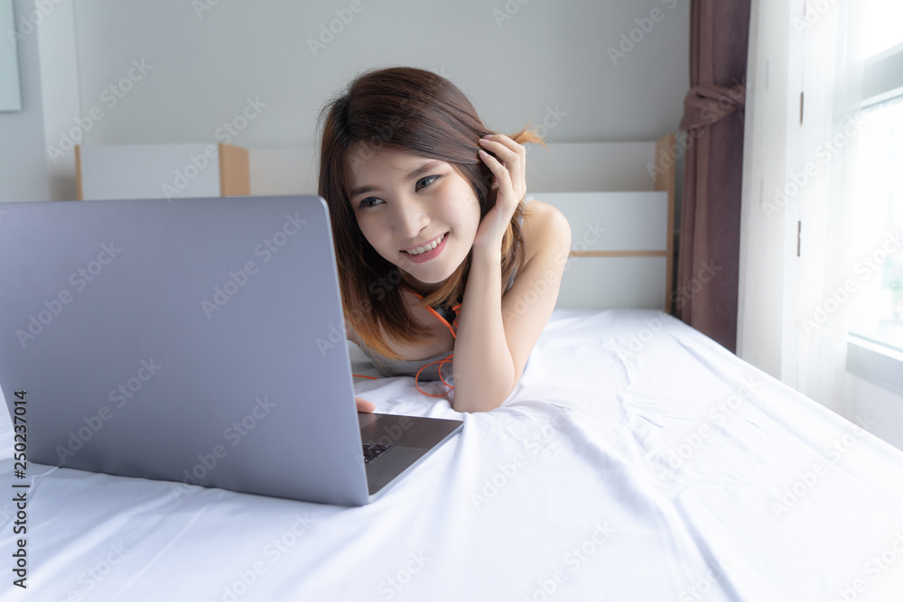 A smiling woman lying down on bed with laptop..Young woman laying on a bed , using and typing on laptop keyboard..Asian girl relaxing in bed on lazy day. Lifestyle education concept. A smiling woman lying down on bed with laptop..Young woman laying on a bed , using and typing on laptop keyboard..Asian girl relaxing in bed on lazy day. Lifestyle education concept.