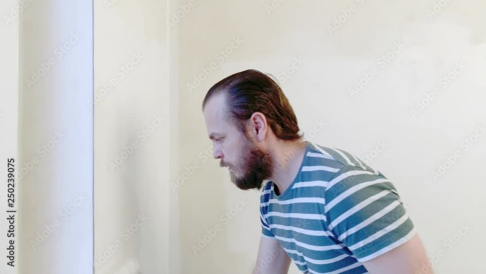 Bearded Man Painting interior Walls at Flat using Paint Roller. Handsome young man doing Repairs - actively paints wall with white paint. Home renovation or Redecoration concept. Bearded Man Painting interior Walls at Flat using Paint Roller. Handsome young man doing Repairs - actively paints wall with white paint. Home renovation or Redecoration concept.