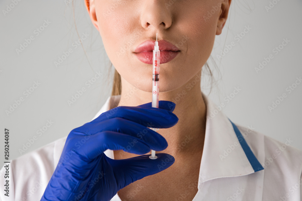 Beautiful young woman cosmetologist doctor isolated over grey wall background holding injection syringe. Beautiful young woman cosmetologist doctor isolated over grey wall background holding injection syringe.