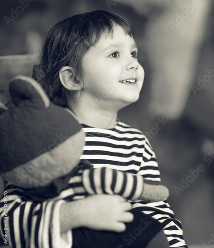 A little happy smiling girl in a skirt and striped t-shirt sitting on a chair and holding an old toy, black and white
