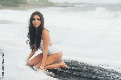 Beautiful smiling girl in white swimwear siting on black sand beach in waves of ocean