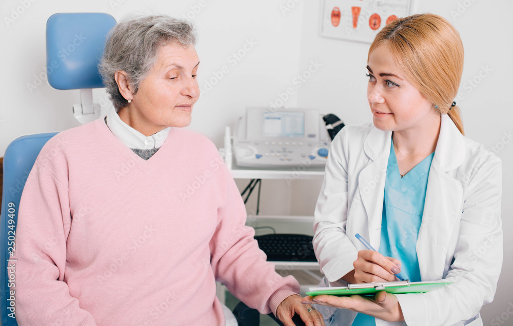 Fototapeta premium Friendly doctor consulting elderly patient in clinic and recording to medical chart