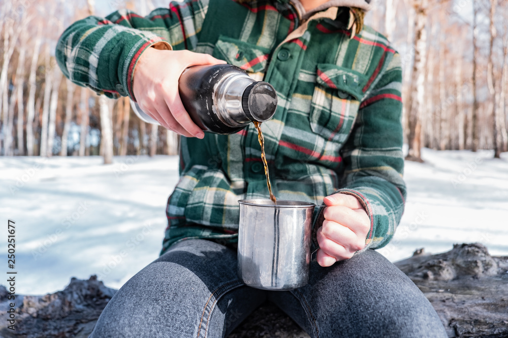 Pouring hot drink out of thermos at a campsite. Person in a winter forest during a hiking trip getting warm Pouring hot drink out of thermos at a campsite. Person in a winter forest during a hiking trip getting warm