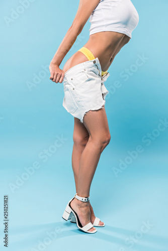 Partial view of graceful woman in yellow panties taking off shorts on blue background
