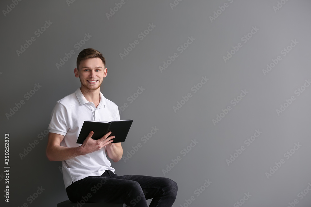 Handsome young man reading book while sitting near grey wall Handsome young man reading book while sitting near grey wall