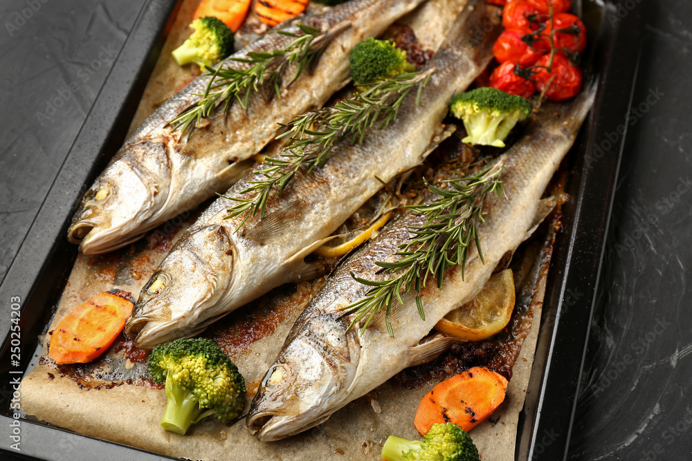 Baking sheet with tasty cooked seabass fish and vegetables on dark table Baking sheet with tasty cooked seabass fish and vegetables on dark table