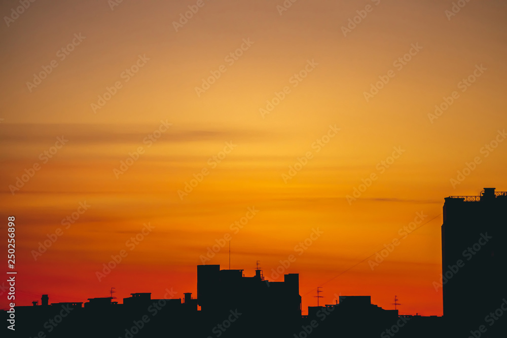 Cityscape with vivid fiery dawn. Amazing warm dramatic cloudy sky above dark silhouettes of city building roofs. Orange red sunlight. Atmospheric background of sunrise in overcast weather. Copy space.