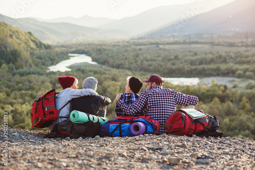 Group of young friends traveling together in mountains. Happy hipster travelers with backpacks sitting on the top of mountain at sunset background. Traveling, tourism and friendship concept.