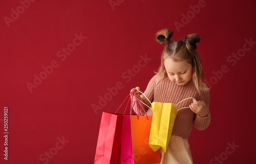Cute little girl looking inside shopping bags on color background