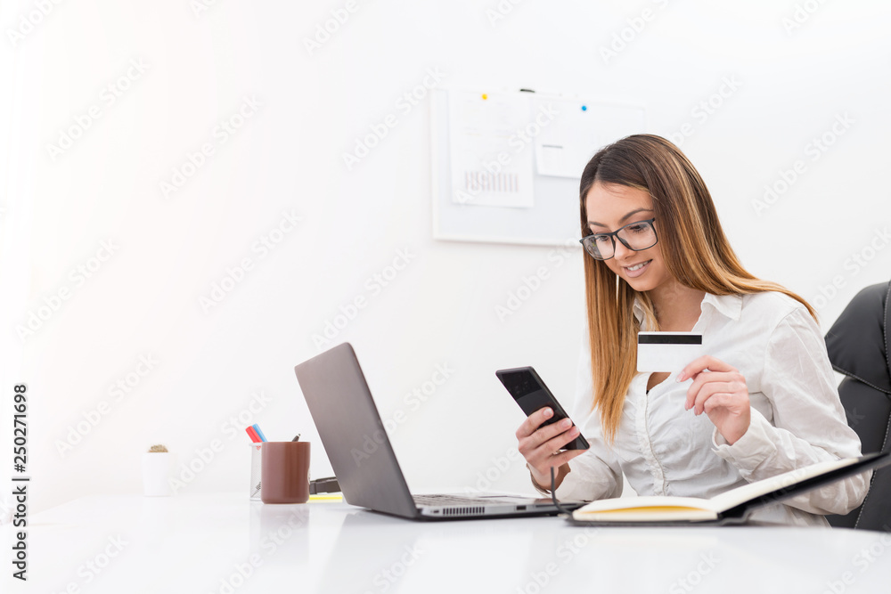 Attractive young businesswoman buying with a bank card on mobile phone while sitting in her office. Attractive young businesswoman buying with a bank card on mobile phone while sitting in her office.