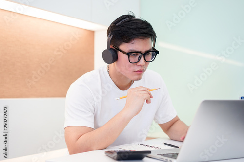 close up young asian student man wearing headset and looking at laptop display for learning mathematics online course from education website , writing lecture on notebook and solve lesson at library 