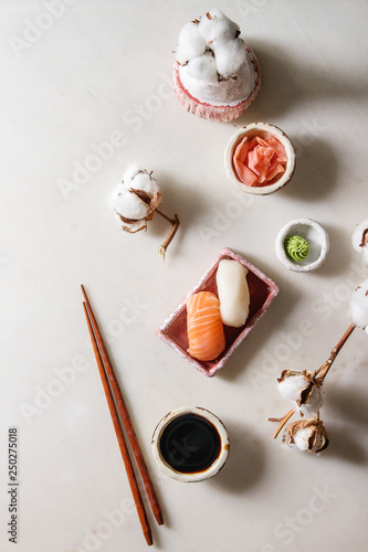 Sushi Set nigiri with salmon and butterfish in pink ceramic serving plate, chopsticks, bowls of soy sauce, and pickled ginger, cotton flowers over white marble background. Flat lay, space. Japan menu