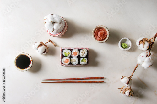 Sushi rolls set in pink ceramic serving plate with chopsticks, bowls of soy sauce, and pickled ginger, cotton flowers over white marble background. Flat lay, space. Japan menu
