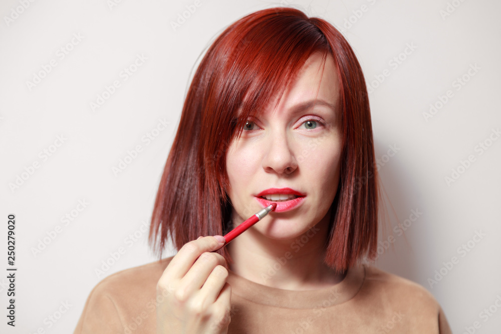 Closeup portrait redhead pretty girl paints her lips with red pencil lipstick. Concept school of makeup, future makeup artist, get ready for party