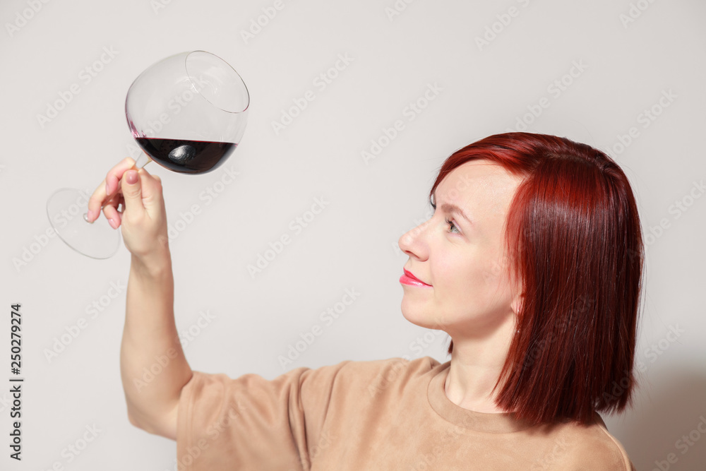 Young beautiful red haired girl in beige dress with bright lipstick on white background smiles, holds glass of red wine by leg assesses color at professional blind tasting sommelier competition Young beautiful red haired girl in beige dress with bright lipstick on white background smiles, holds glass of red wine by leg assesses color at professional blind tasting sommelier competition