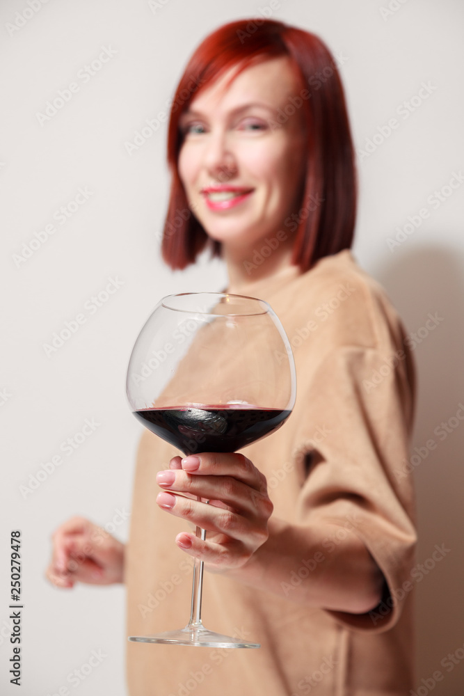 Young beautiful redhaired woman with bright lipstick on white background is smiling and holding a glass of red wine at professional blind tasting sommelier competition Young beautiful redhaired woman with bright lipstick on white background is smiling and holding a glass of red wine at professional blind tasting sommelier competition