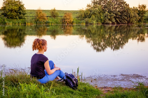 A young girl sits on the riverbank and contemplates the beauty of the surrounding nature_