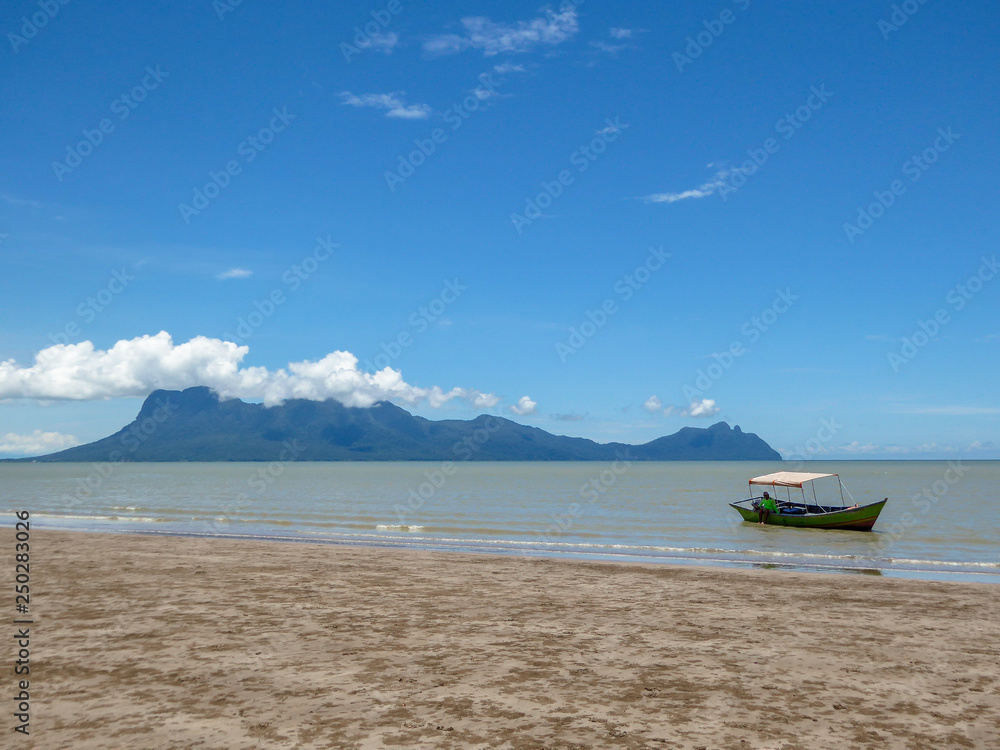 Fototapeta premium A beach in Bako National Park in Borneo, Malaysia. Yellow sand mixing with algae on the ground. Boat parked on the shore of the beach. High mountains in the back, covered with white clouds.