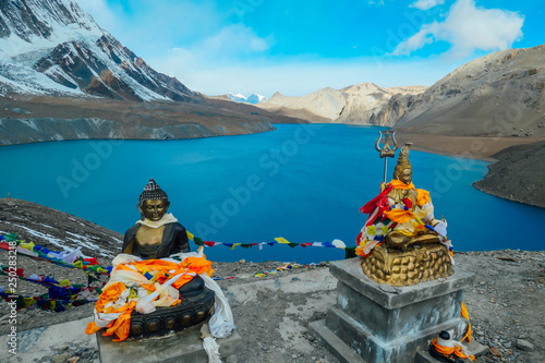 Two Buddha statues at the Tilicho Lake, covered with prayer's flags. Blue and calm surface of the lake, mountains covered in the shadow, sunlight in the back. Annapurna Circuit Trek, Nepal. photo