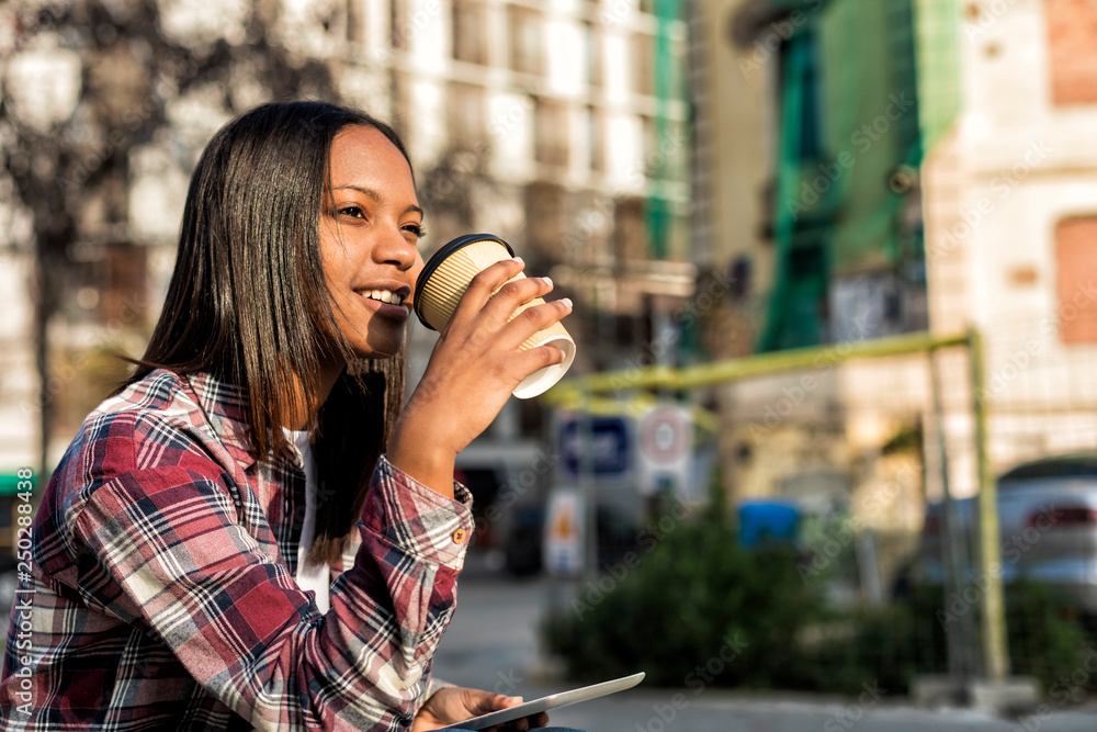 Young beautiful african american woman sitting outdoors in the city with a tablet pc while holding a take away coffee in a sunny day