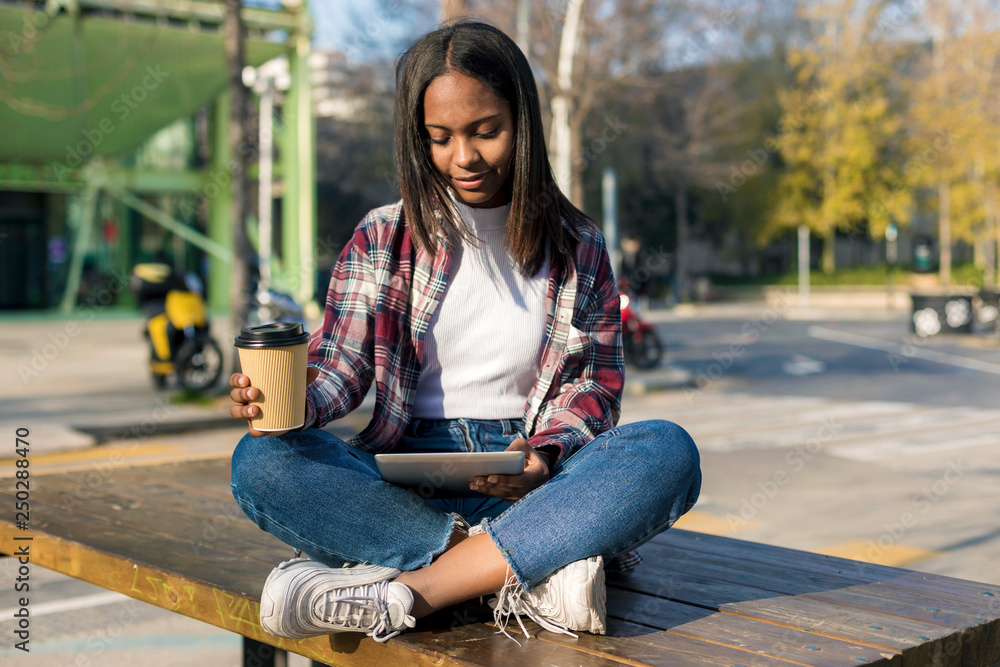 Young beautiful african american woman sitting outdoors in the city with a tablet pc while holding a take away coffee in a sunny day - obrazy, fototapety, plakaty 