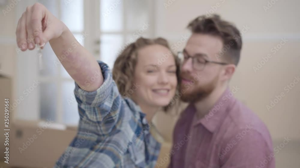 Portrait of a couple in love, they just bought a house and moving. Woman extends a hand with keys to the camera, man kissing her in cheek. Focus changes from keys to people faces Portrait of a couple in love, they just bought a house and moving. Woman extends a hand with keys to the camera, man kissing her in cheek. Focus changes from keys to people faces