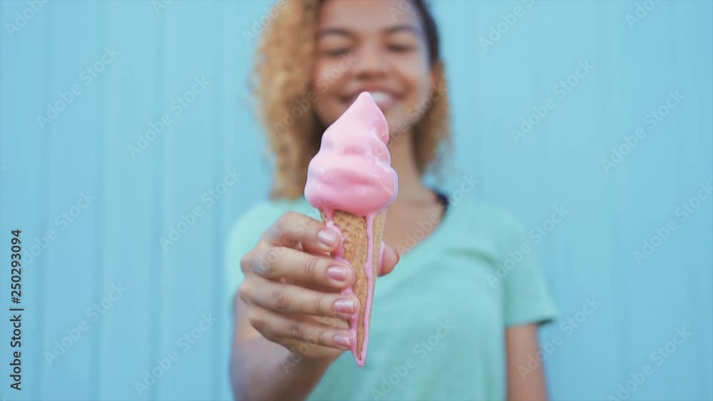 young afro girl eating pink melting ice cream at blue wall background young afro girl eating pink melting ice cream at blue wall background