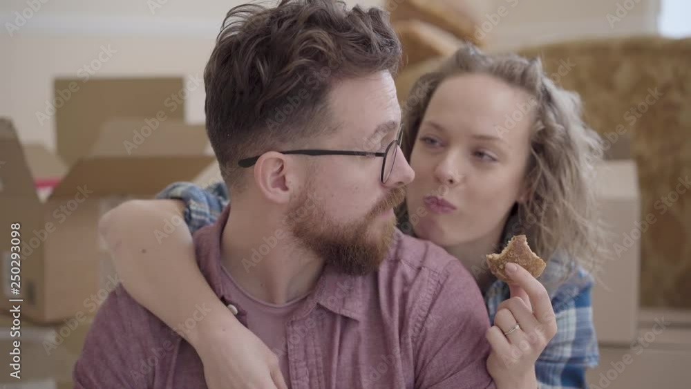 Portrait of pretty woman eats cookie, hugging from behind bearded man in glasses close up. Unpacked boxes are in the background. Married couple moves into a new home. Family rest together Portrait of pretty woman eats cookie, hugging from behind bearded man in glasses close up. Unpacked boxes are in the background. Married couple moves into a new home. Family rest together