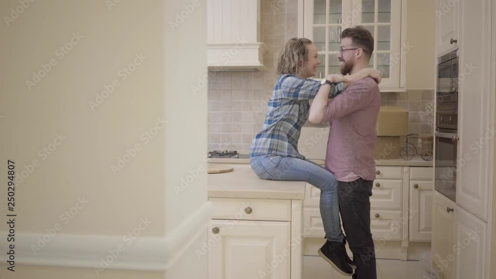 Bearded man stands near pretty woman sitting on table in big half empty kitchen. Woman hugs her husband, they kissing ans smiling. Married couple moves into a new home. Camera moves left behind wall Bearded man stands near pretty woman sitting on table in big half empty kitchen. Woman hugs her husband, they kissing ans smiling. Married couple moves into a new home. Camera moves left behind wall