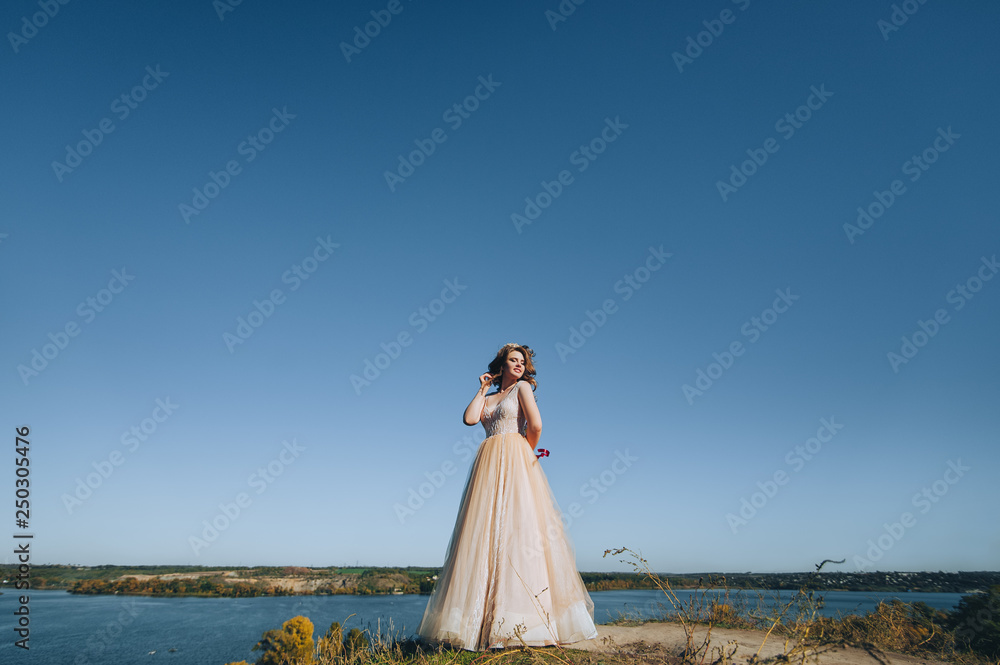 A lovely bride in a beige dress with a crown is standing on a cliff, against a background of rocks and a blue sky. Wedding photography. Princess Bride. A lovely bride in a beige dress with a crown is standing on a cliff, against a background of rocks and a blue sky. Wedding photography. Princess Bride.