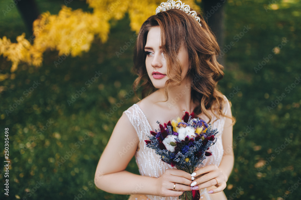 Fototapeta premium A smiling bride in a beige dress and a bouquet is standing in a park with yellow leaves. The princess with a crown enjoys the fall. Portrait of a beautiful bride with brown hair. Wedding photography.