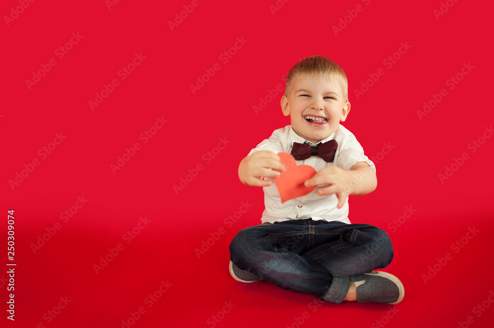 for holidays, lovers day and mothers day, baby boy sits on a red background with a red heart in his hands. The concept is to give gifts for holidays, lovers day and mothers day, baby boy sits on a red background with a red heart in his hands. The concept is to give gifts