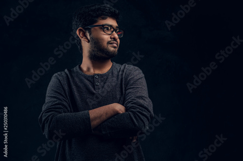 Studio portrait of a young Indian guy wearing eyewear and casual clothes posing with his arms crossed and looking sideways in a dark room
