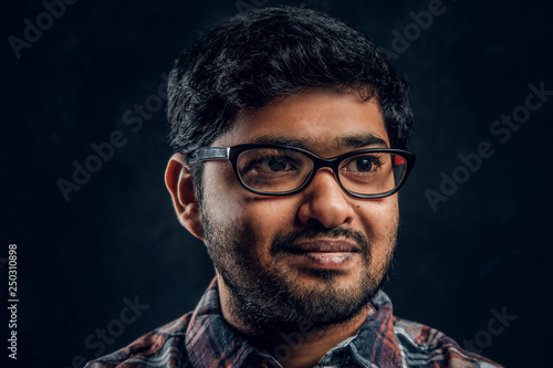 Close-up portrait of a young Indian guy in eyewear and checkered shirt in studio against the background of the dark wall