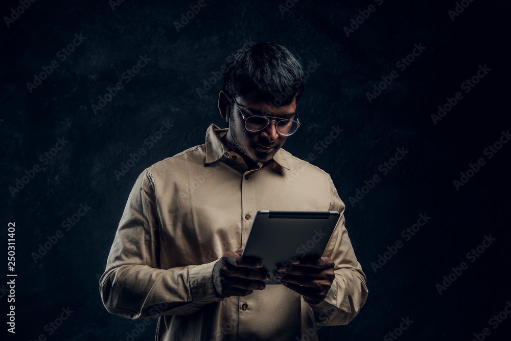 Indian male in eyewear and shirt using a tablet computer in studio against the background of the dark wall Indian male in eyewear and shirt using a tablet computer in studio against the background of the dark wall