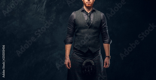 Handsome redhead man dressed in an elegant vest with tie and kilt in studio against a dark textured wall