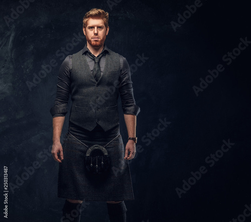 Handsome redhead man dressed in an elegant vest with tie and kilt in studio against a dark textured wall
