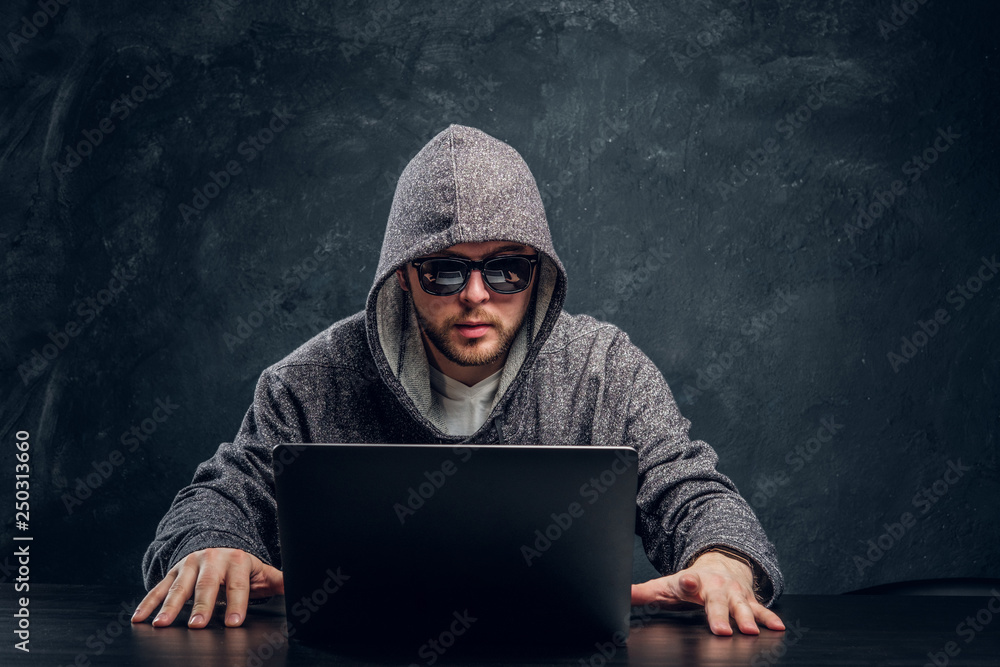 Programmer in hoodie and sunglasses hacking passwords on laptop sitting at a desk in a dark office