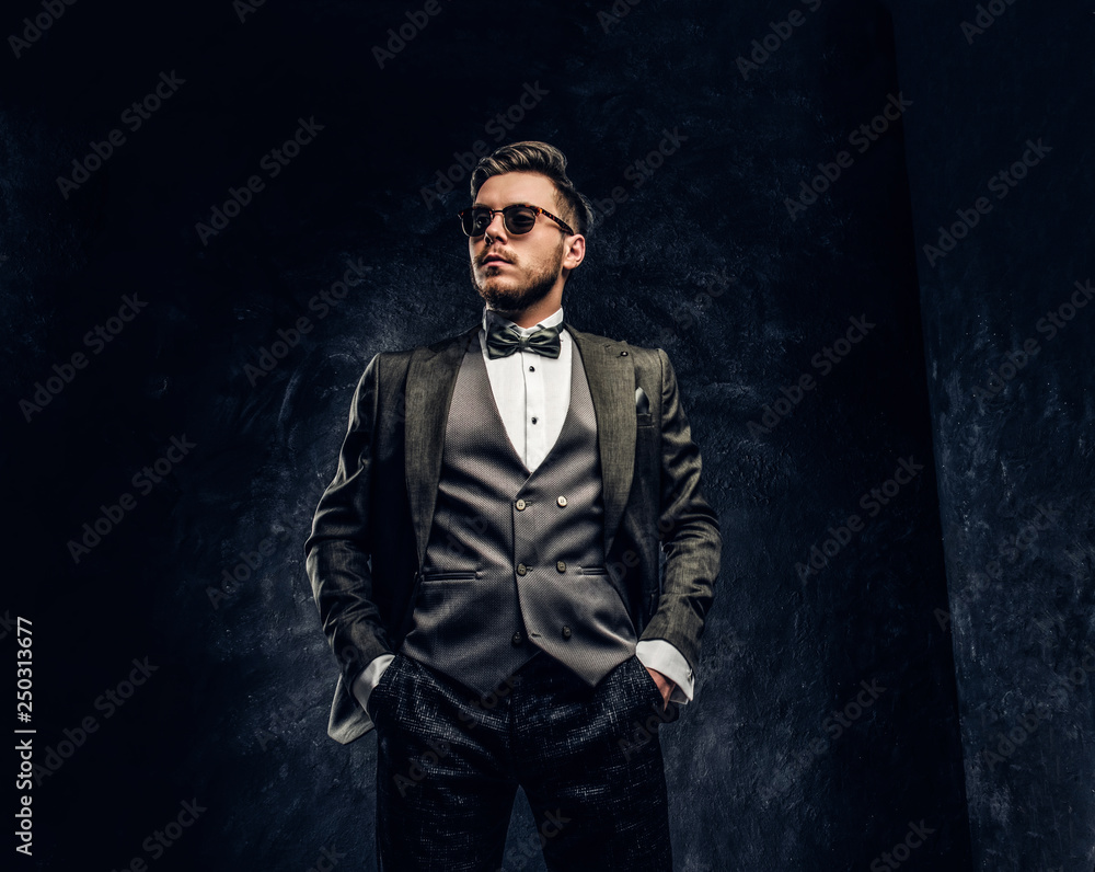 Obraz premium A stylish young man in sunglasses dressed in an elegant suit posing with a hand in a pocket against a dark textured wall