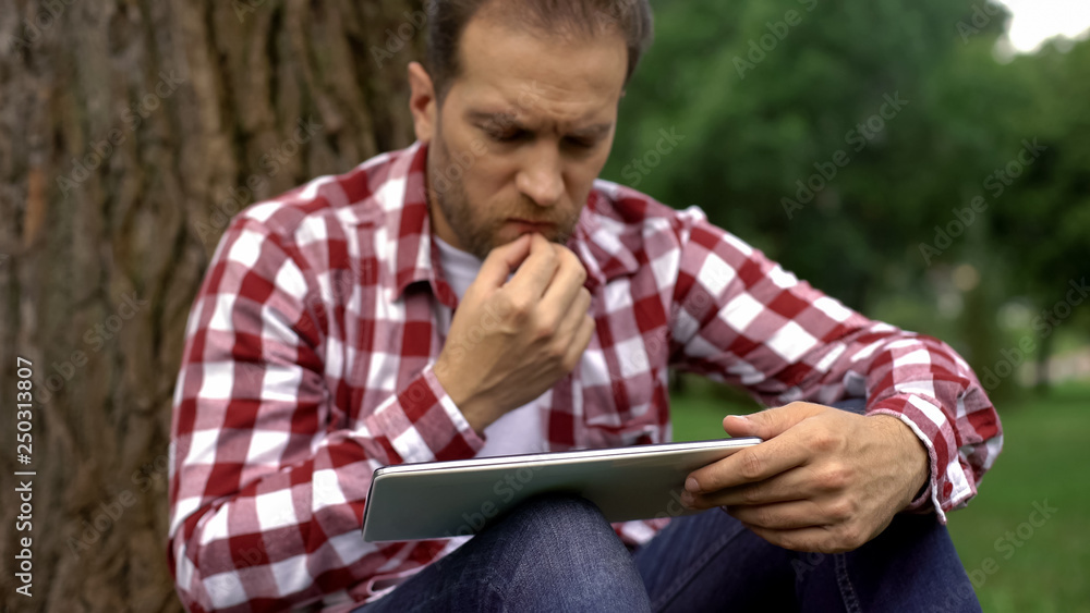 Concentrated adult men using tablet in park, reading article about mens health Concentrated adult men using tablet in park, reading article about mens health