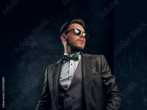 A stylish young man in sunglasses dressed in an elegant suit posing with a hand in a pocket against a dark textured wall