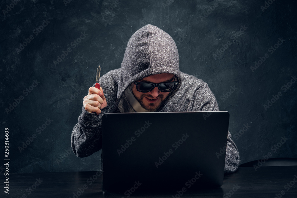 Hacker in hoodie and sunglasses holds a knife lock-up sitting behind a  laptop in a dark room Stock Photo | Adobe Stock