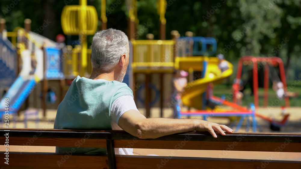 Male pensioner sitting on bench and watching grandkids on playground, family Male pensioner sitting on bench and watching grandkids on playground, family